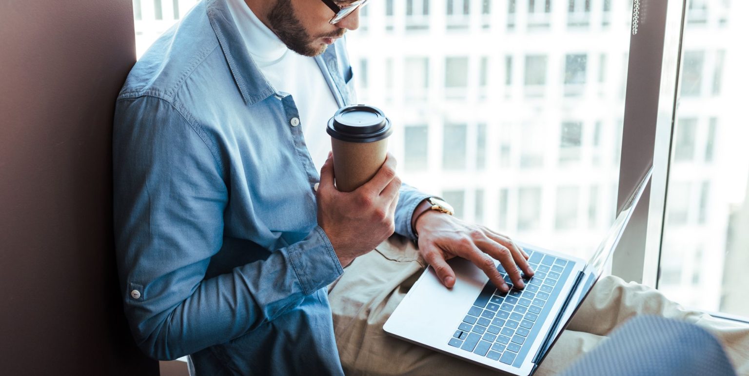 Mand i blå skjorte drikker kaffe og arbejder på bærbar computer